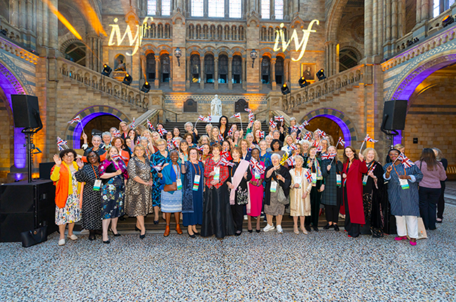 Group of diverse women in colorful outfits pose together in a historic stone hall, waving Union Jack flags at a conference or event.