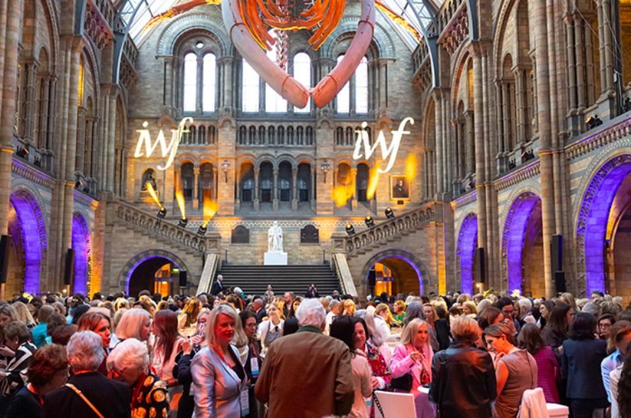 Crowd of people mingling at a formal reception inside a grand historic hall with arched windows and neon 'iwf' signs overhead.