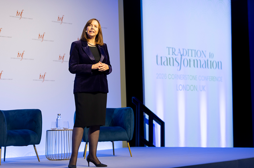 Woman in a dark blazer and black dress standing on a conference stage, with blue chairs and a small table beside her; backdrop reads 'Tradition to Transformation' and 'London, UK'.