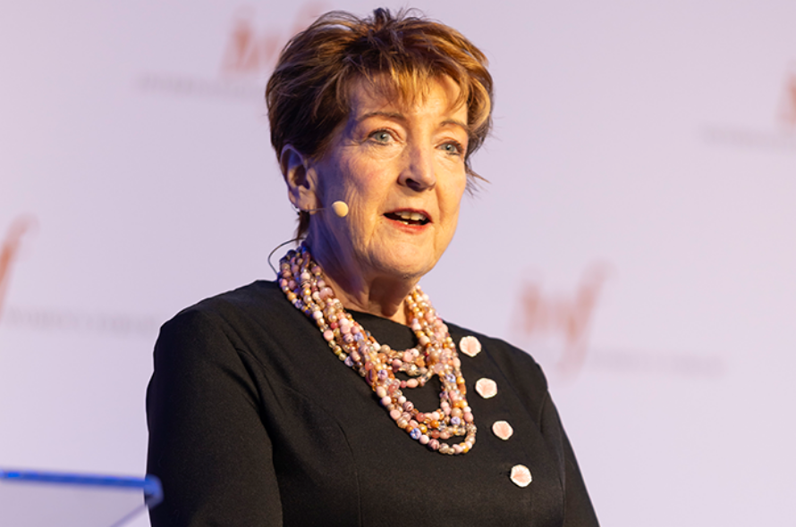 Older woman wearing a black dress and pink pearl necklace speaking on stage with a headset microphone.