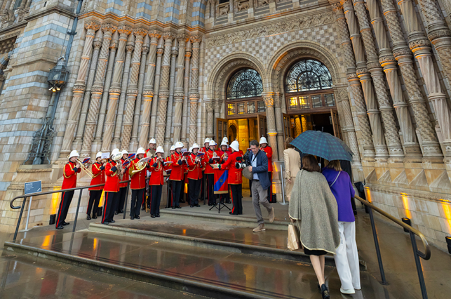 A red-uniform marching band with white helmets performs on the steps of an ornate stone building as a conductor directs, with spectators watching nearby and one person holding an umbrella.