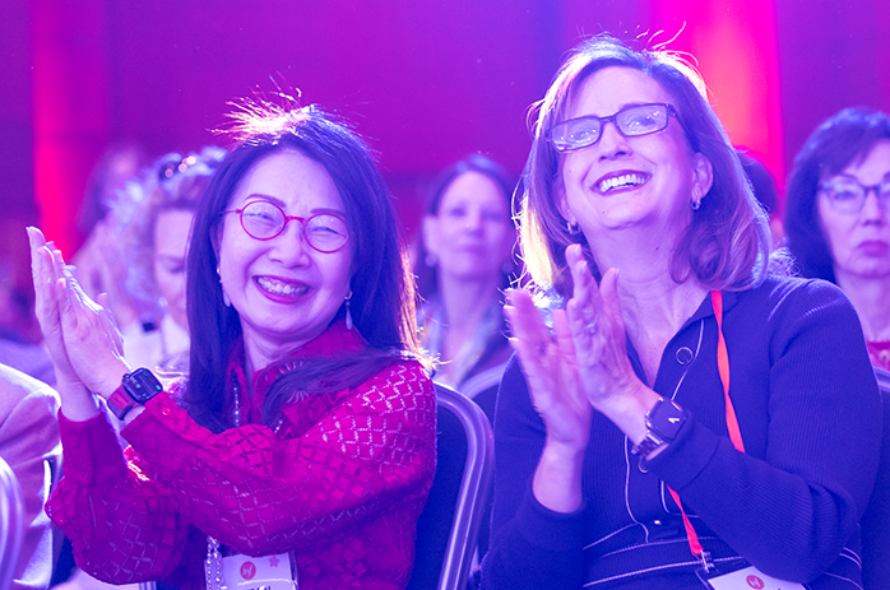 Two smiling women in the audience clap at a conference, one in a red lace top with pink glasses and badges visible.
